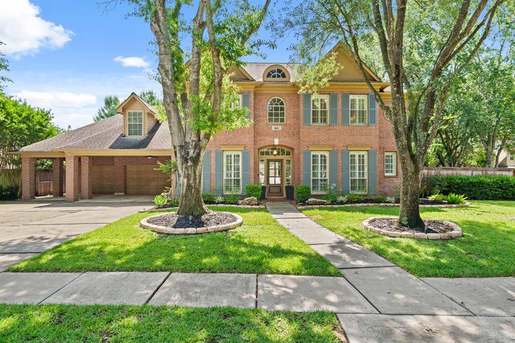 This beautiful two-story brick home features a welcoming front entrance with large windows and shutters. It has a spacious driveway leading to a three-car garage, surrounded by a well-maintained lawn and mature trees, providing a charming curb appeal.