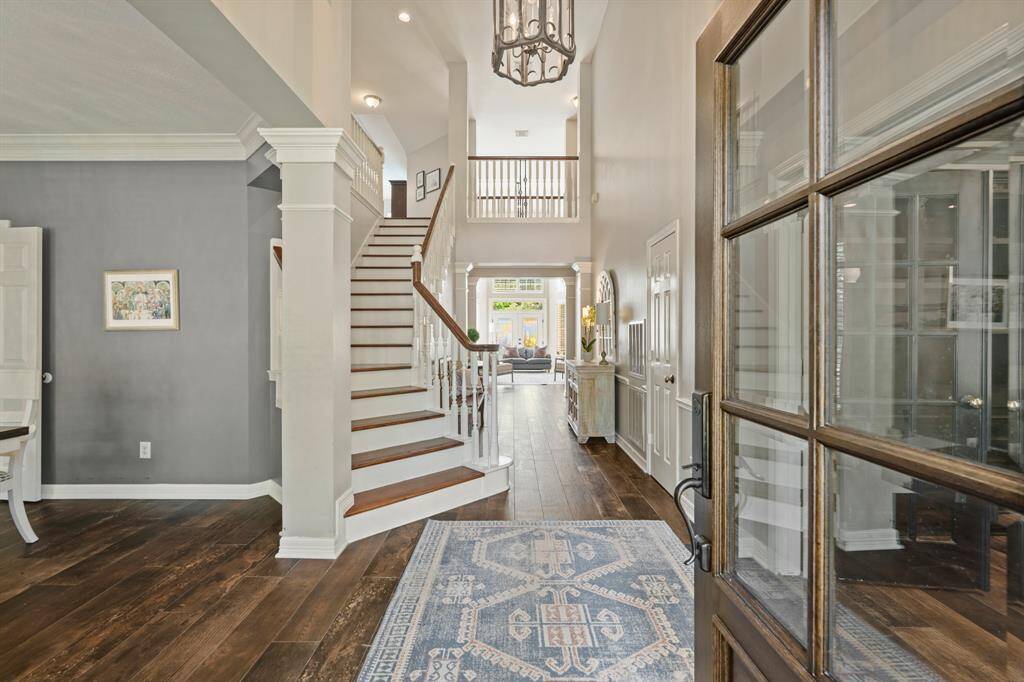 This welcoming entryway features a high ceiling with elegant lighting, an open staircase with wood accents, and wood stairway flooring. The space is bright and inviting, leading into a living area with a view to the outdoor POOL/SPA oasis.
