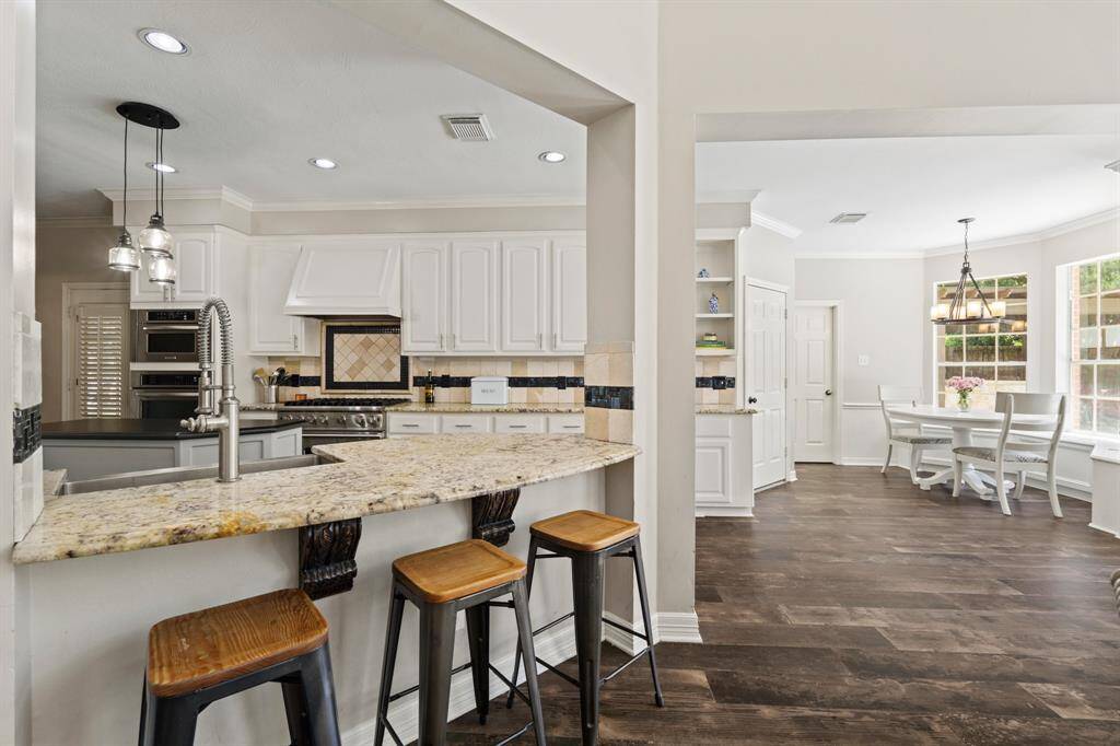 This photo showcases a bright, open-concept kitchen and dining area. The kitchen features granite countertops, a breakfast bar with seating, pendant lighting, and white cabinets. The adjacent breakfast room space has large windows, window box seating, natural light and a cozy atmosphere.