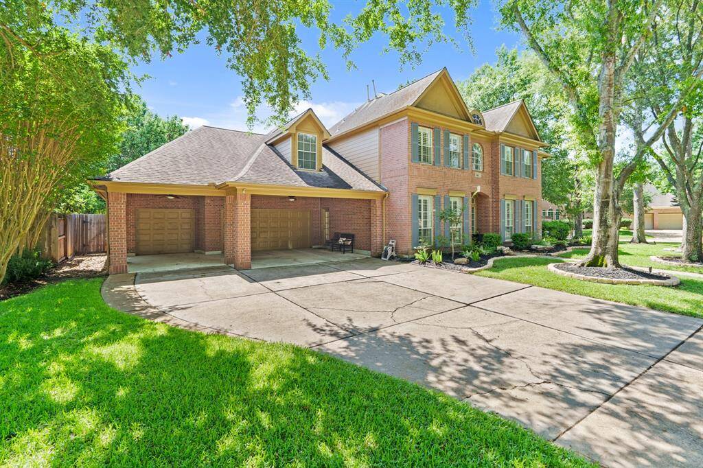 This photo shows a spacious two-story brick home with a three-car garage and additional parking underneath the porte-cochere with easy access door to kitchen.. It features a well-maintained driveway and lush, green landscaping with mature trees.