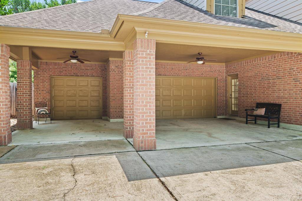 This photo features a spacious three-car garage with a porte-cochere, red brick exterior, and ceiling fans. There’s a small seating area to the side entrance, creating a welcoming and functional entrance.