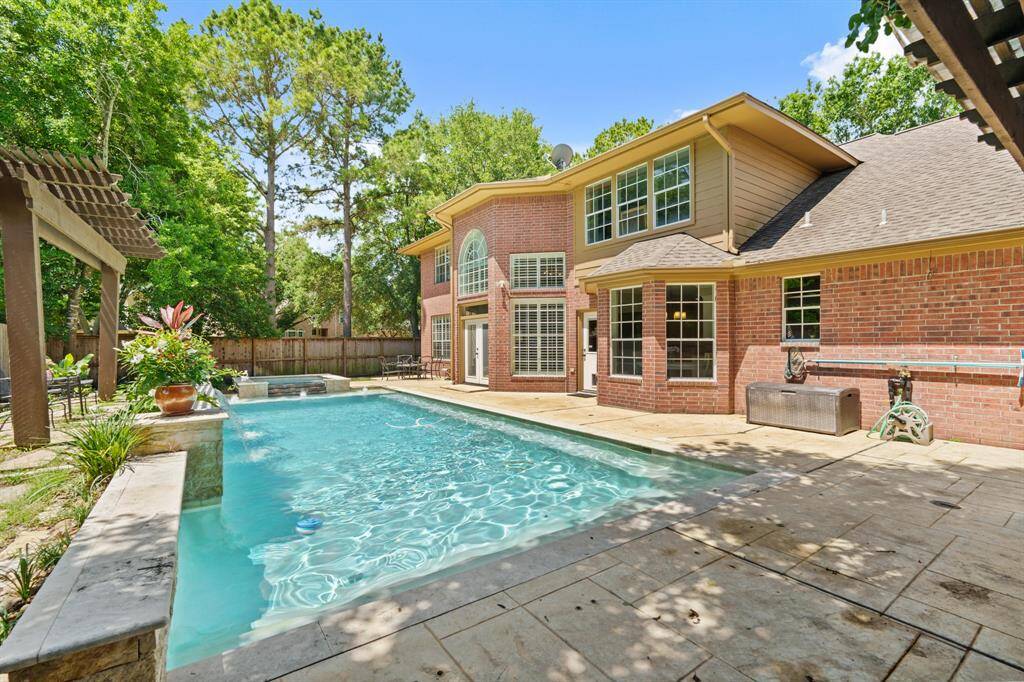 This photo showcases the backyard with a large Swimming POOL/SPA. The area is surrounded by mature trees, providing privacy. A PERGOLA covered patio with built-in grill, mini fridge, sink and bar area adds to the outdoor living space, great for entertaining.