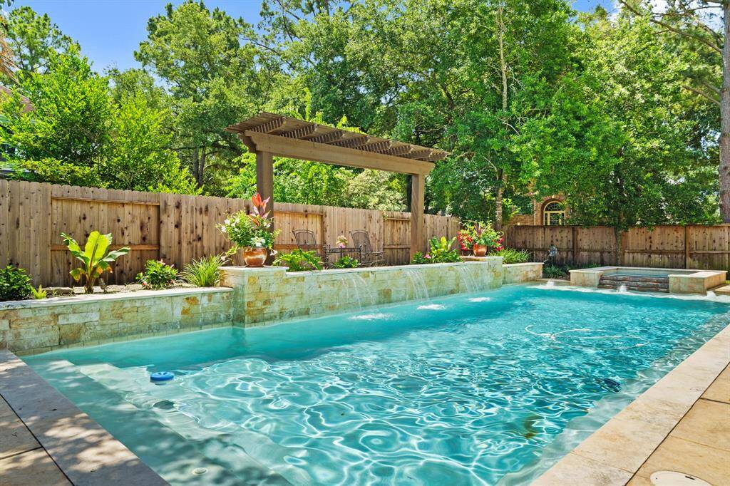 This backyard features a luxurious POOL/SPA with a waterfall, surrounded by lush greenery and a wooden privacy fence. There's a pergola for shade, creating a serene and private oasis.