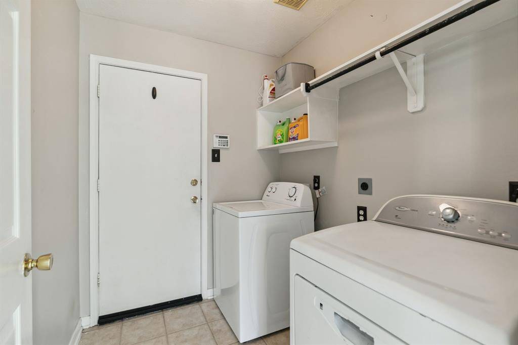 This laundry room features a compact yet efficient layout, complete with a washer and dryer, overhead shelving for convenient storage, and direct access to the outdoors. Neutral-toned walls and durable tiled flooring combine practicality with understated style.