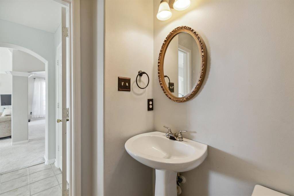 A compact, well-lit bathroom featuring a pedestal sink, a round mirror with decorative trim, and an overhead light fixture. Neutral wall tones and a glimpse into the adjoining bright living area enhance the sense of openness and continuity