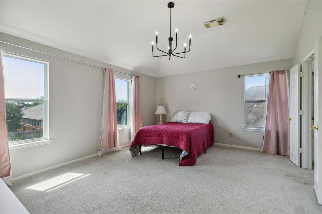 This expansive bedroom is bathed in natural light from large windows, accentuated by a vaulted ceiling and a striking modern chandelier. Neutral carpeting adds warmth underfoot, while soft pink curtains lend a touch of elegance and charm, creating a serene and inviting retreat.