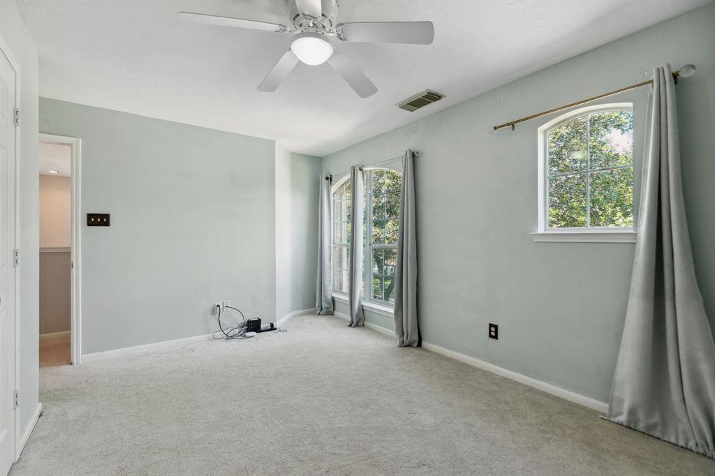 This front room features soft green walls, carpeted floors, and ample natural light from two large windows. It includes a ceiling fan, silver curtains, and offers a spacious, airy feel.