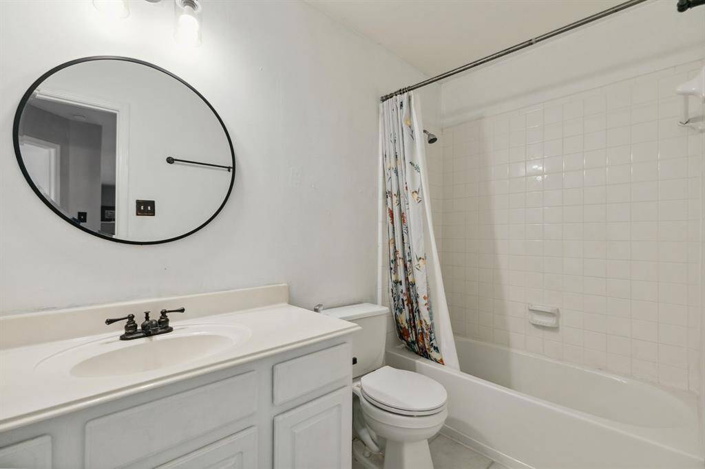 This 2nd bathroom features a clean, modern design with a white vanity and a circular mirror. It includes a tiled shower-tub combo with a decorative shower curtain, offering a simple yet functional space.