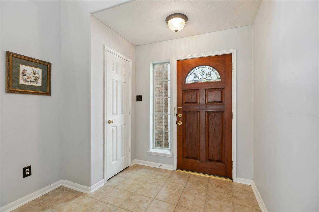 This welcoming entryway features a beautifully crafted wooden front door with a decorative glass panel, tiled flooring, and a carpeted staircase leading to the upper level. Large windows provide natural light, creating a bright and inviting atmosphere.