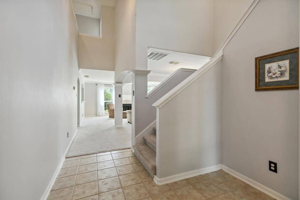 Walk into this open entryway with elegant tiled flooring that transitions seamlessly into a cozy, carpeted living area. A staircase to the right adds architectural interest, while light-colored walls and soaring ceilings enhance the sense of openness. Natural light pours in through a window at the back, creating a bright and airy atmosphere."