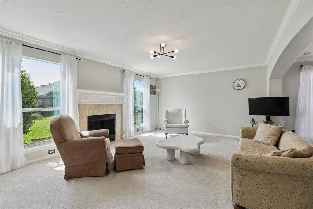 This expansive living room showcases a soothing neutral palette, centered around a cozy fireplace that adds warmth and charm. Large windows flood the space with natural light, highlighting the plush carpeting and elegant chandelier above. An arched entryway seamlessly connects the living area to the kitchen, enhancing the open-concept design and inviting easy flow throughout the home.
