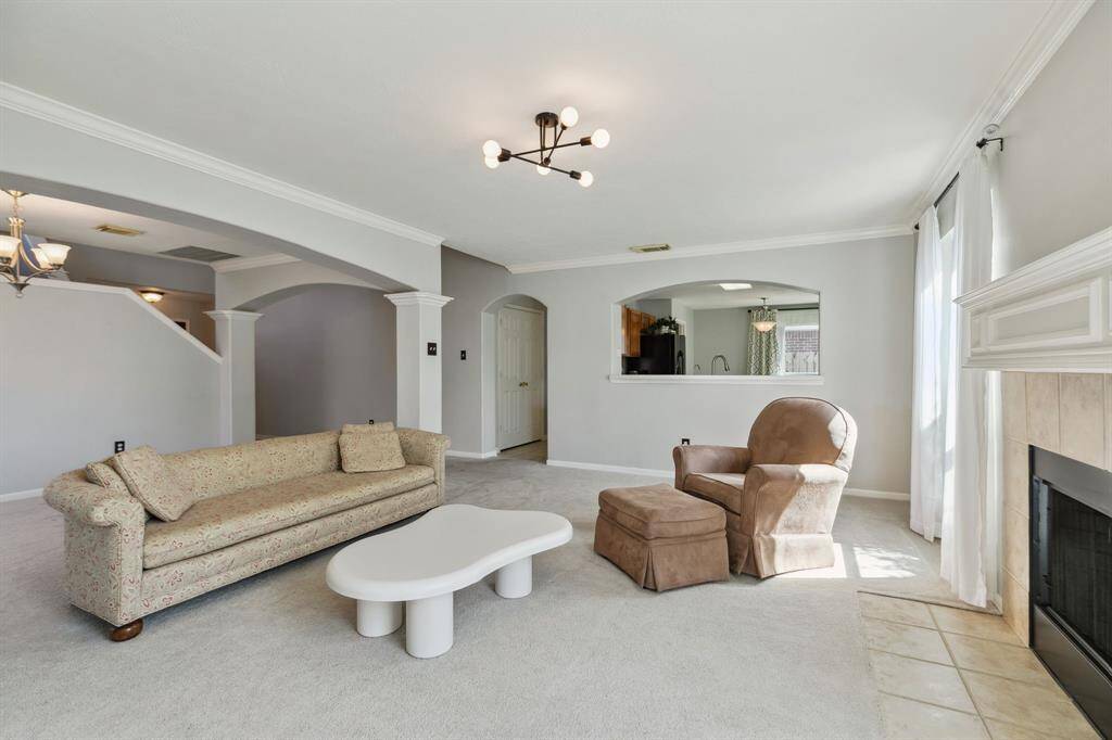 This expansive living room showcases a soothing neutral palette, centered around a cozy fireplace that adds warmth and charm. Large windows flood the space with natural light, highlighting the plush carpeting and elegant chandelier above. An arched entryway seamlessly connects the living area to the kitchen, enhancing the open-concept design and inviting easy flow throughout the home.