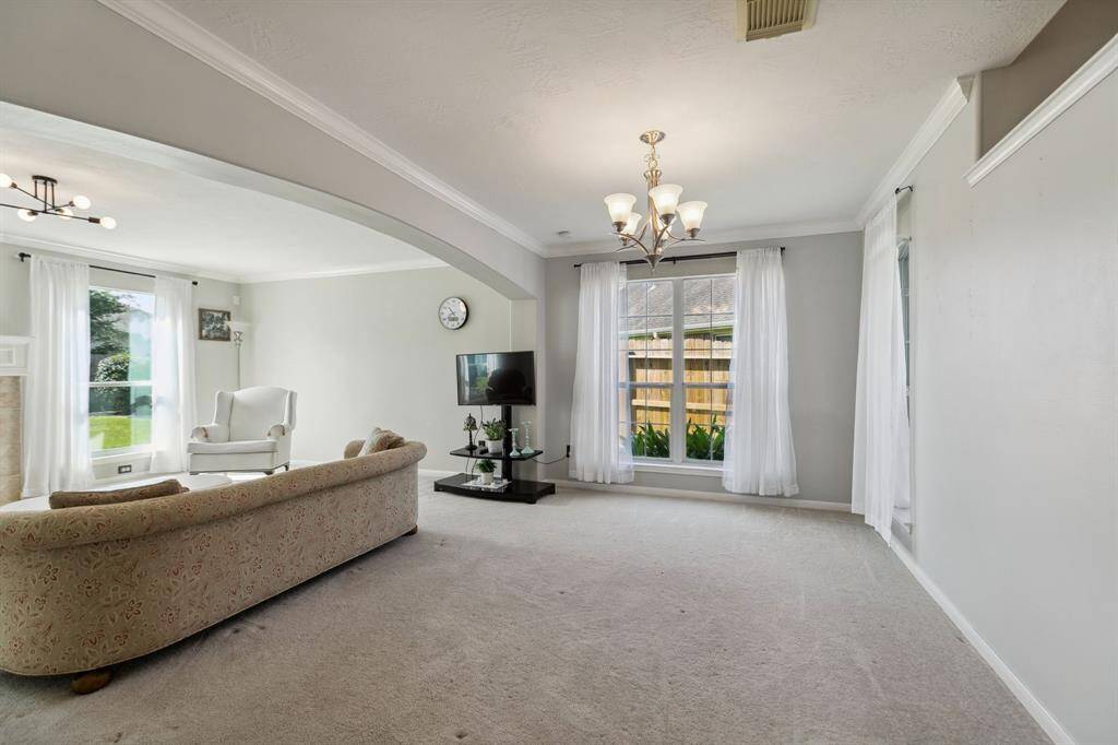 Bright and inviting dining room with neutral walls and carpeting. Features large windows with sheer curtains for natural light, a cozy seating area, and modern light fixtures. Open layout offers a spacious feel.