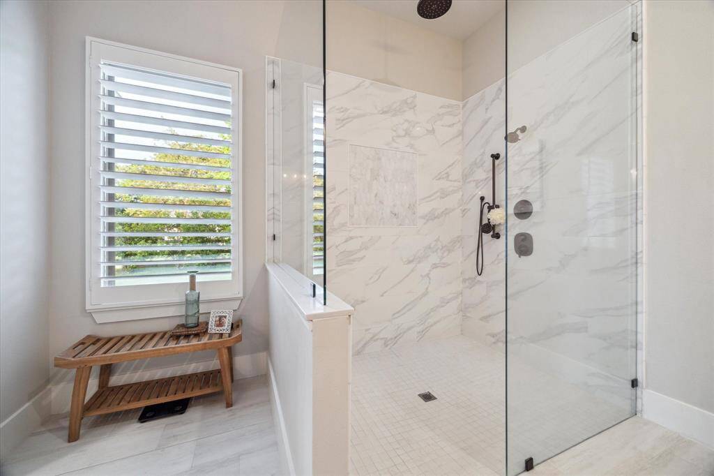 Spa-style walk-in shower with marble tile, rainfall head, and a serene sitting area beside sunlit windows.