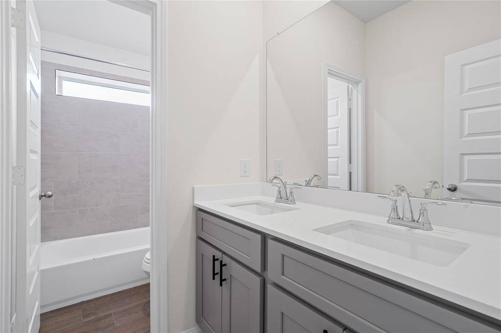 Secondary bath features wood look tile flooring, bath/shower combo with tile surround, stained wood cabinets, beautiful light countertops with dual vanities, mirror, sleek fixtures and modern finishes.