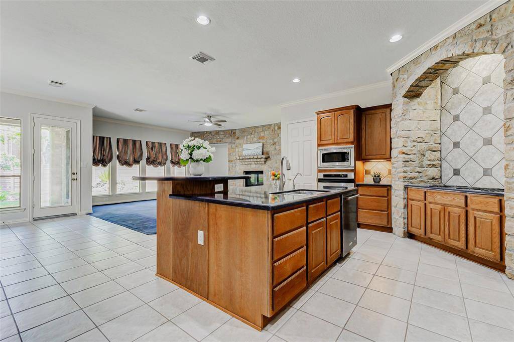 This spacious kitchen features elegant stone accents, stainless steel appliances, and ample wooden cabinetry. It has a large island with a sink, tile flooring, and plenty of natural light from large windows and a glass door leading outside. Perfect for entertaining and family gatherings.