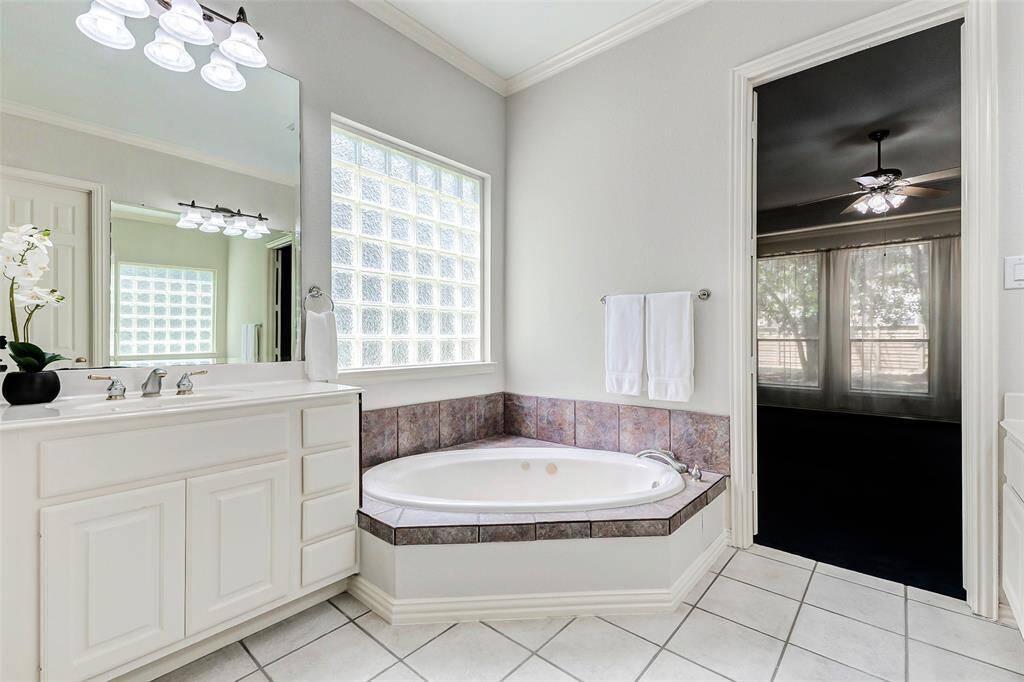 The primary bathroom features a spacious corner jetted bathtub with tile accents, a large vanity and ample natural light from a glass block window providing privacy. The adjoining primary bedroom is visible through an open door.