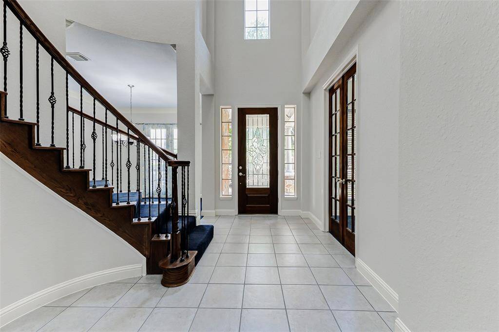 The bright entryway features high ceilings and tiled flooring. The elegant staircase features dark wood and wrought iron railings. Glass-paneled double doors lead to an adjacent room, and the front door has decorative glass inserts, allowing natural light to fill the space.