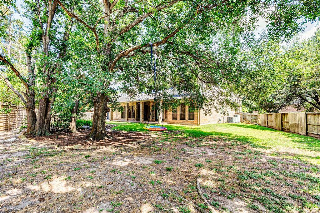 A spacious backyard with mature trees providing ample shade, a swing for enjoyment, and a fenced perimeter for privacy. The house features a covered patio perfect for outdoor relaxation. On the side are the 3 A/C units for the home.
