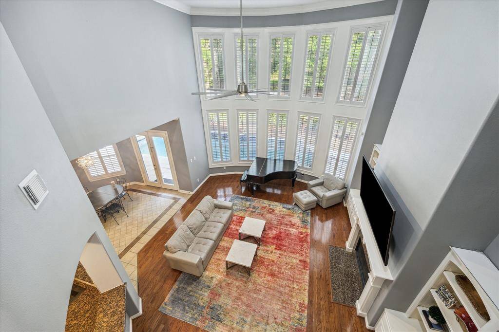 Bird’s Eye View of Everyday Elegance: From the upstairs landing, take in the two-story living room framed by plantation shutters, extensive crown molding, and a wall of windows that flood the home with natural light and showcase the backyard retreat.
