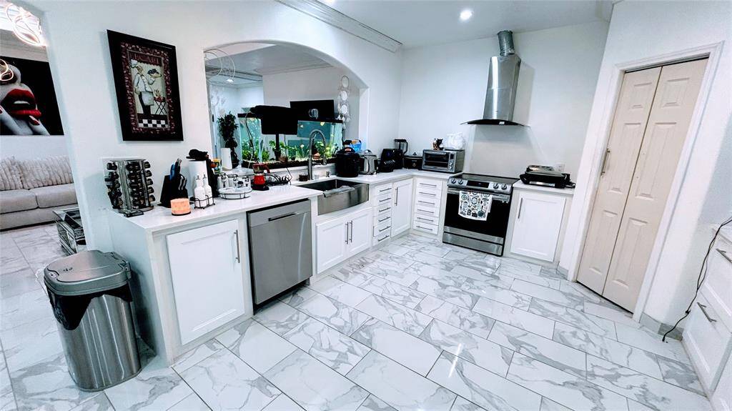 This kitchen features modern white cabinetry, stainless steel appliances, and elegant marble-patterned tile flooring. It includes a sleek range hood, ample counter space, and an open layout with a view into the adjacent living area.