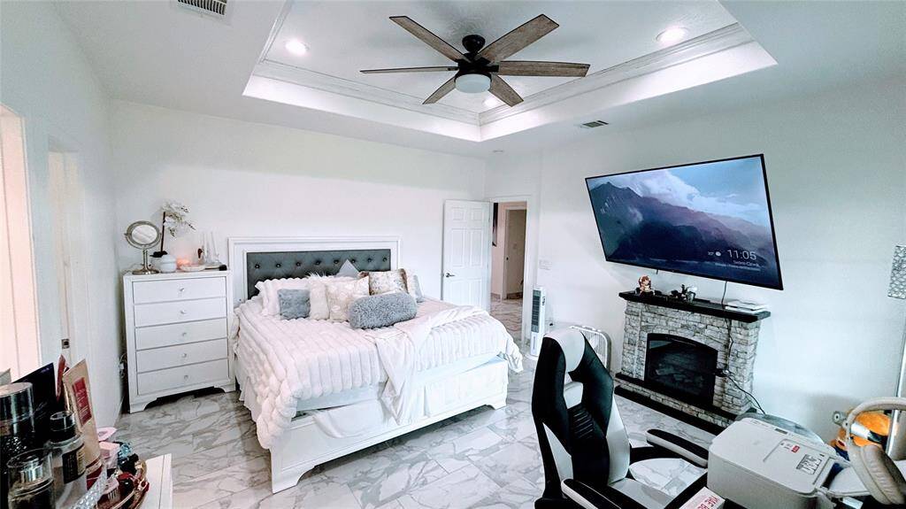 This spacious Master bedroom features a cozy fireplace, large mounted TV, and a ceiling fan. It has a modern aesthetic with a tray ceiling, marble-like flooring, and ample natural light.