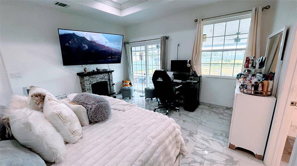This spacious Master bedroom features a cozy fireplace, large mounted TV, and a ceiling fan. It has a modern aesthetic with a tray ceiling, marble-like flooring, and ample natural light.