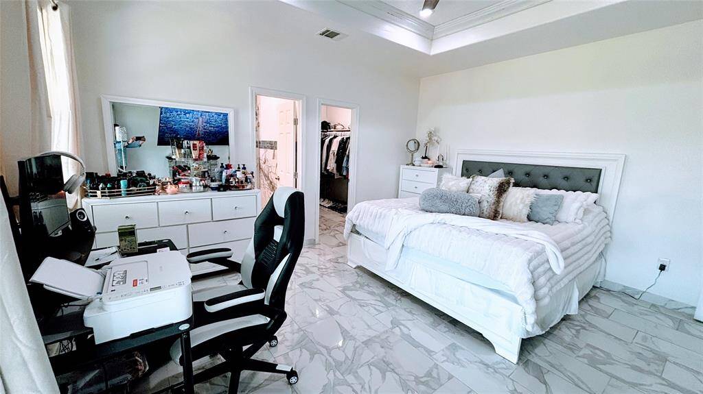 This spacious Master bedroom features a cozy fireplace, large mounted TV, and a ceiling fan. It has a modern aesthetic with a tray ceiling, marble-like flooring, and ample natural light.