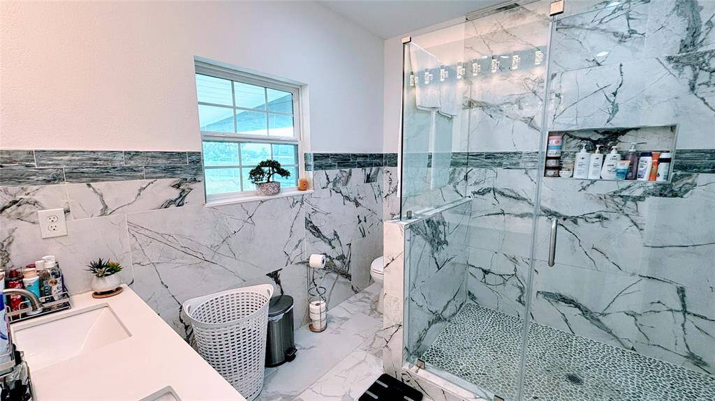 This modern master  bathroom features elegant marble-patterned tiles, a spacious glass-enclosed shower, and ample natural light from a well-placed window. The design includes a sleek vanity area and practical shelving, making it both stylish and functional.