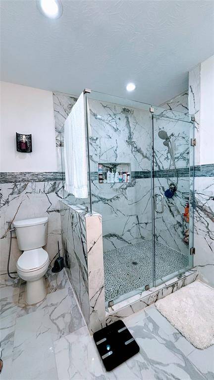 This master bathroom features a modern design with elegant marble tile throughout. It includes a glass-enclosed shower with a pebble floor, a built-in niche for toiletries, and a standard toilet.
