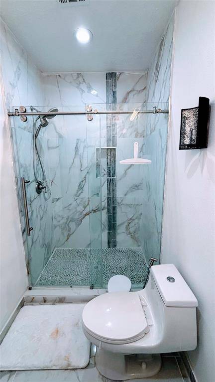This modern bathroom features a sleek, glass-enclosed shower with marble-patterned walls and a pebble stone floor, complemented by a contemporary toilet and bright, clean finishes.