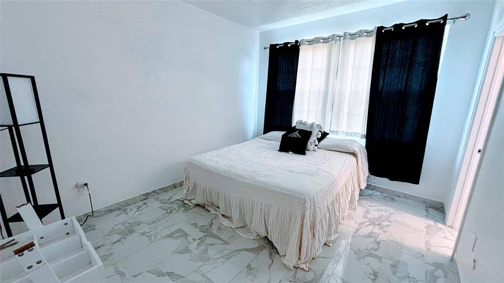 This bright bedroom features marble-patterned tile flooring, a cozy bed with elegant bedding, and ample natural light from the large windows.