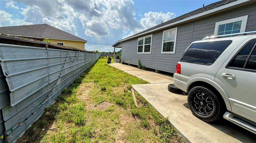 The side of a house with gray siding, featuring a concrete driveway and a grassy area alongside a metal fence.