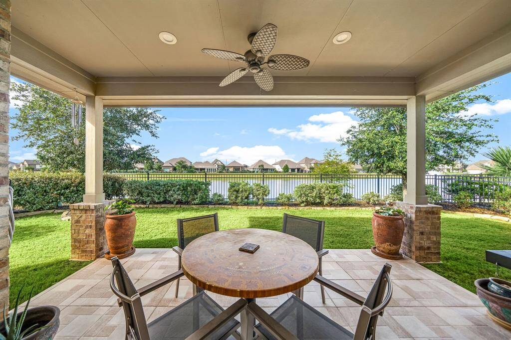Enjoy tranquil waterfront living from this covered patio oasis, showcasing a serene lake view framed by lush landscaping and elegant brick columns. The spacious outdoor retreat features a stylish tile floor, ceiling fan, and recessed lighting, creating the perfect setting for relaxation or entertaining—all just steps from a beautifully manicured lawn.