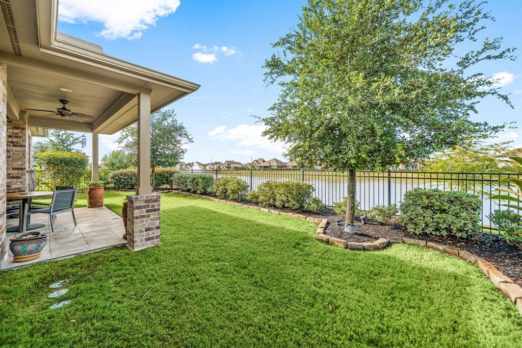 Step into a peaceful backyard escape where a covered patio overlooks a scenic lake, framed by well-maintained greenery and classic brick pillars. The space is enhanced with sleek tile flooring, built-in lighting, and a ceiling fan for year-round comfort—offering the ideal backdrop for quiet mornings or outdoor gatherings.