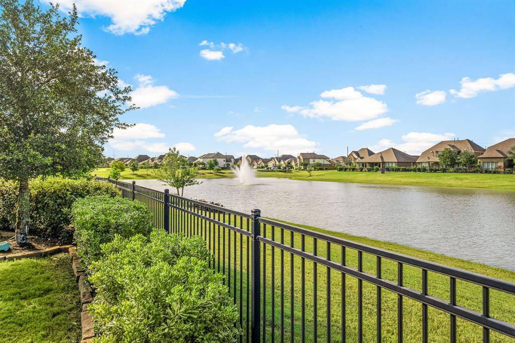 Take in breathtaking waterfront views from this beautifully landscaped backyard, featuring a wrought iron fence that offers unobstructed sightlines to a tranquil lake and elegant fountain. Surrounded by manicured greenery and set beneath open skies, this outdoor space invites peaceful moments and scenic relaxation.