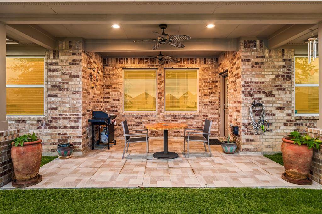 Unwind in this charming covered patio retreat, where warm brick accents and soft lighting create a welcoming ambiance. Enjoy peaceful lake views framed by manicured greenery and a wrought iron fence, all enhanced by the convenience of a built-in grill and ceiling fan for year-round enjoyment.