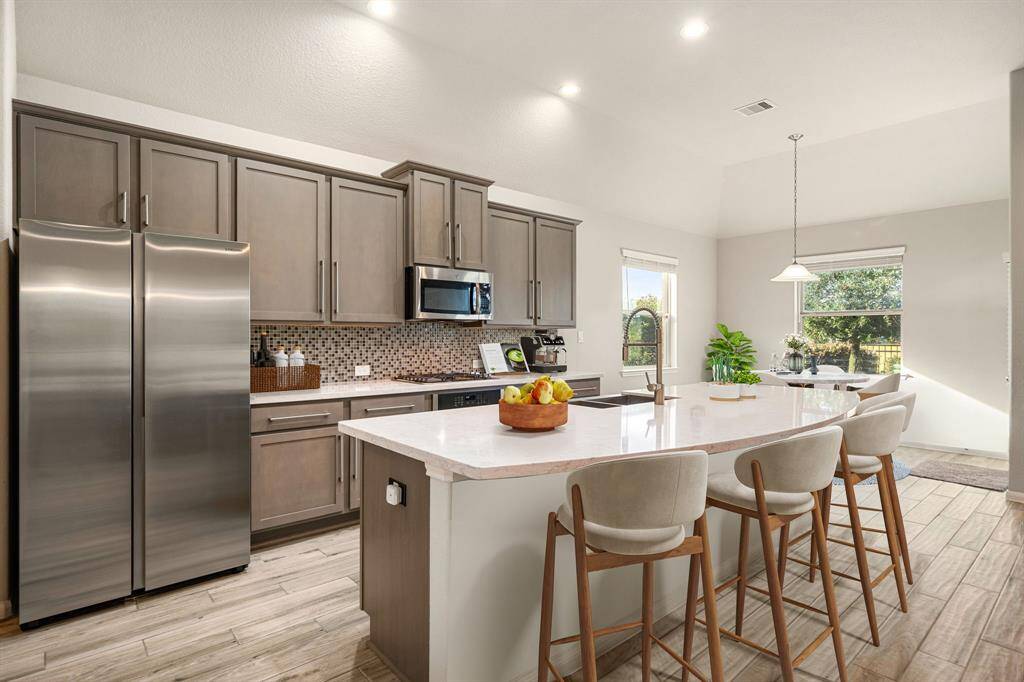 This stunning kitchen features sleek grey cabinetry, Quartz countertops, and a spacious island ideal for meal prep and casual dining. A custom tile backsplash adds elegance, while stainless steel appliances and a deep basin sink enhance functionality. Natural light pours in through large windows, highlighting the open layout and warm wood-look tile flooring. Perfectly designed for both everyday living and entertaining.