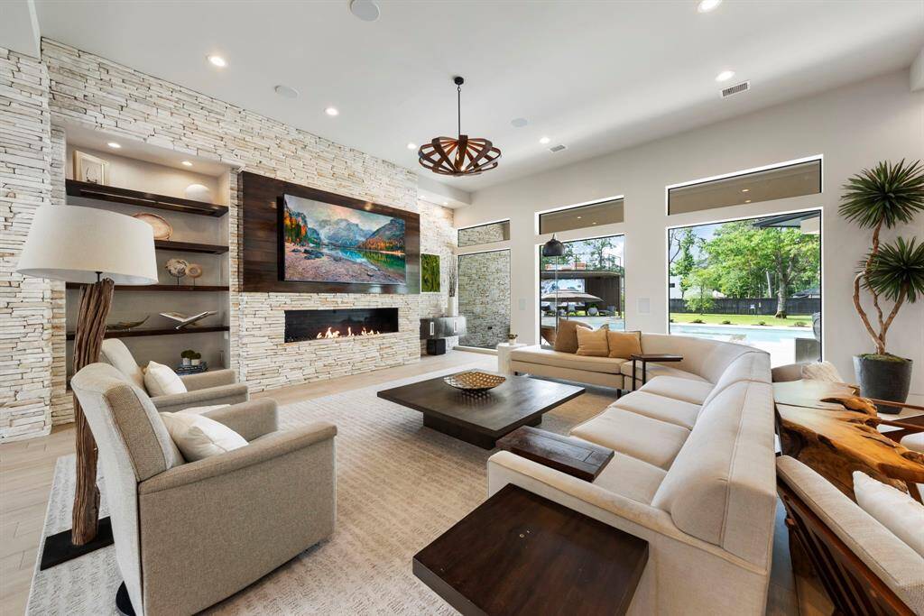 This sun-drenched family room stuns with floor-to-ceiling stonework, a sleek linear fireplace, and expansive windows that frame lush outdoor views like a living painting.