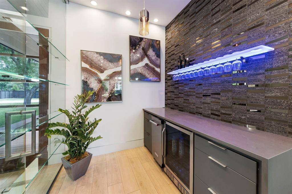 This sleek, glass-enclosed bar showcases a moody tile backsplash and illuminated glassware display with ice maker and beverage fridge.