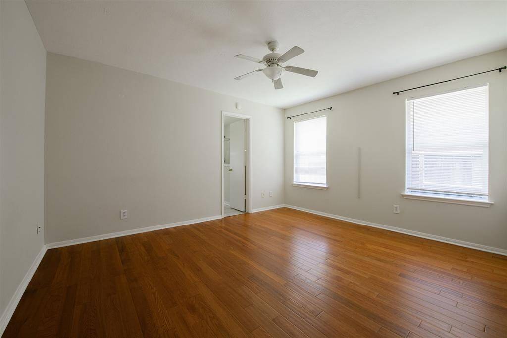 Another angle of the spacious primary suite highlights its open layout and clean finishes. Wood-look floors offer elegance and warmth, while the ceiling fan and large windows ensure a light and airy atmosphere. A peaceful retreat with simple lines and timeless appeal.