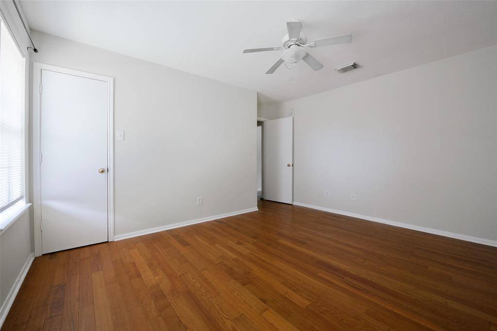 This alternate view of the primary suite showcases its spacious layout, ceiling fan, and natural light. The rich wood-look flooring adds warmth, while neutral paint offers a blank canvas for any design style. Two doorways provide convenient access to closets and the en-suite bath.