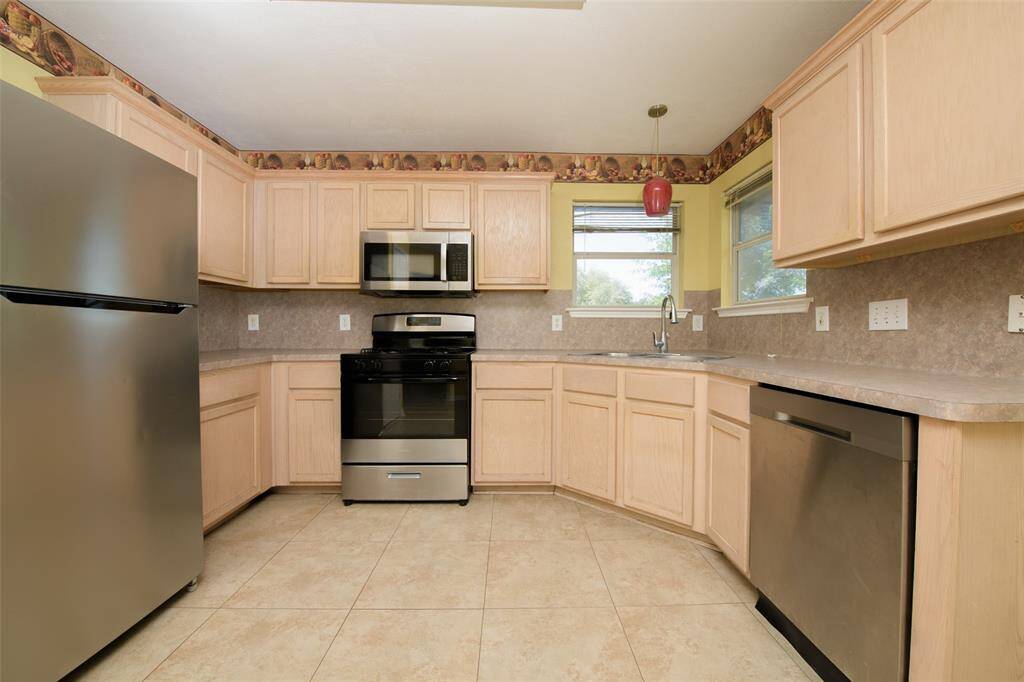 The kitchen features updated stainless steel appliances including a gas range, microwave, dishwasher, and refrigerator. Warm-toned cabinets and neutral countertops offer ample storage and prep space, while windows above the sink provide a view of the backyard.