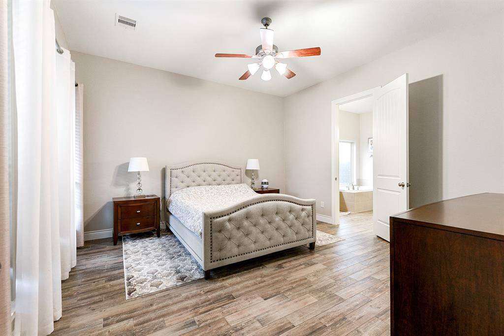The primary en suite features matching wood-look tile flooring, high ceilings with a drop-down ceiling fan, and windows creating a serene and private retreat.