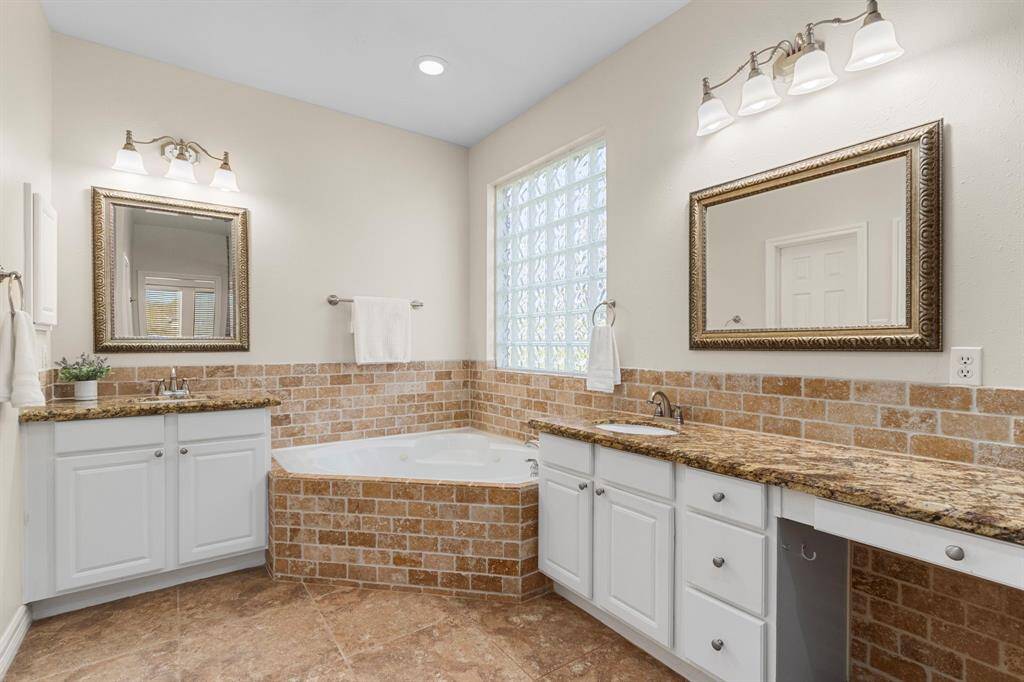 The Primary Bath features elegant 18x18 tile floors, double vanities with granite countertops and a stylish 2x4 brick pattern travertine backsplash. A luxurious jetted tub sits beneath a blocked glass window, providing both natural light and privacy for a relaxing retreat.