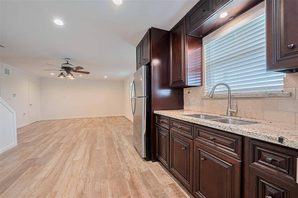 View from the kitchen into the open living area, highlighting the rich cabinetry, dual sink, stainless steel fridge, and continuous wood-look tile flooring under recessed lighting and a ceiling fan.