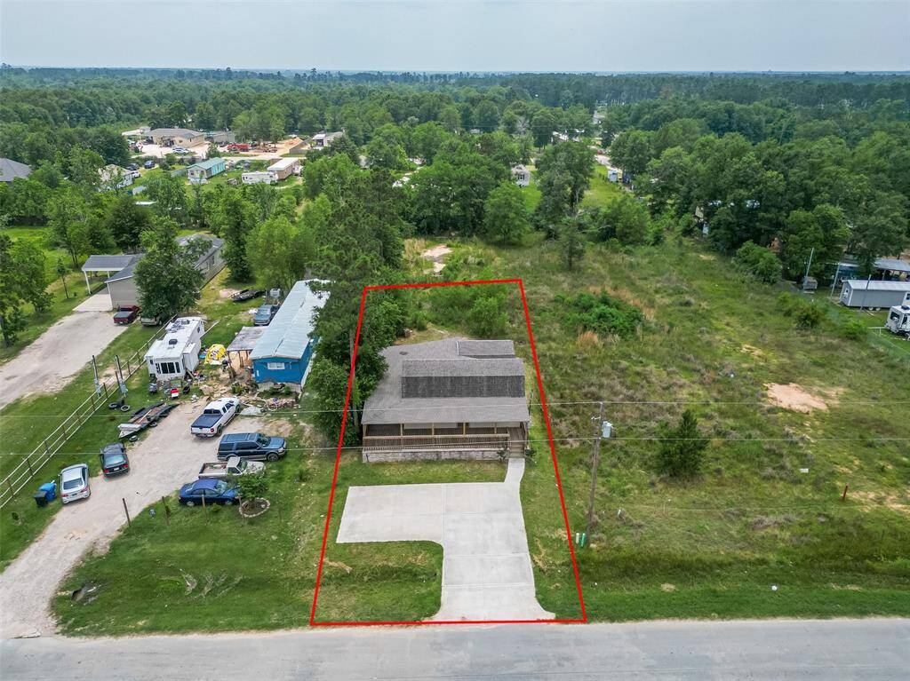 This aerial photo shows a single-family home outlined in red, with a large driveway and wraparound porch. The lot is spacious with trees in the back and open grassy areas. The home is surrounded by a mix of neighboring houses, RVs, and wooded land, giving it a quiet, semi-rural feel.