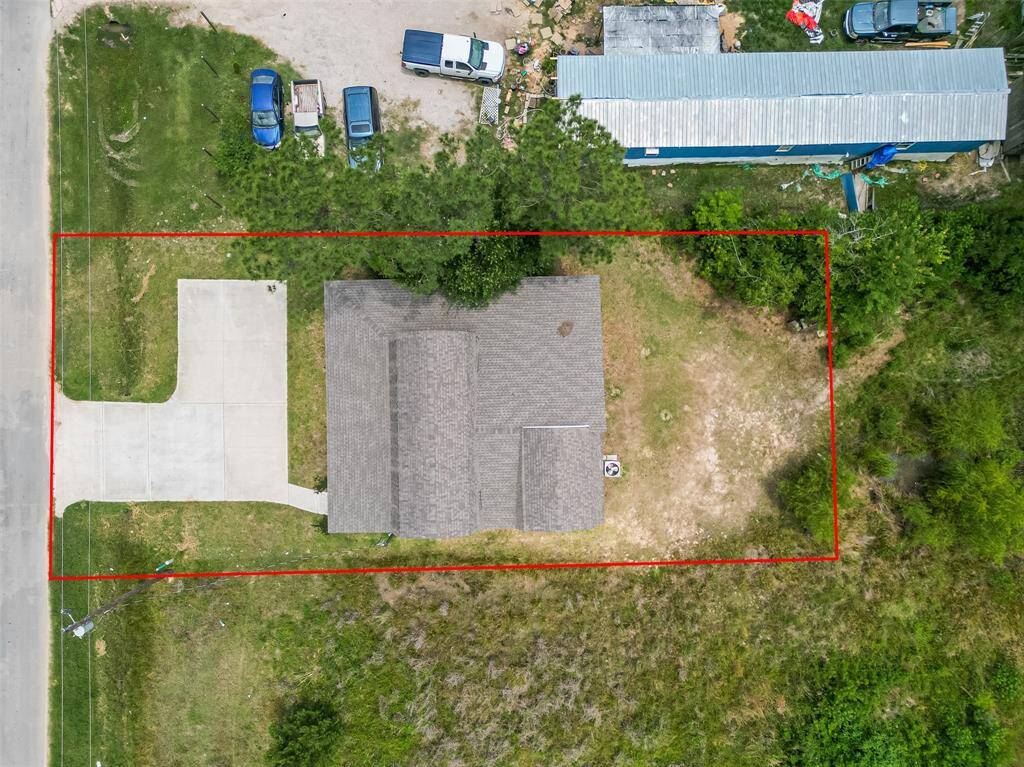 This top-down aerial view shows a single residential property outlined in red. The home sits near the front of the lot with a wide concrete driveway and a wraparound porch. The backyard is spacious and mostly undeveloped, bordered by trees and greenery. The lot is rectangular and flat, offering potential for further landscaping or development.