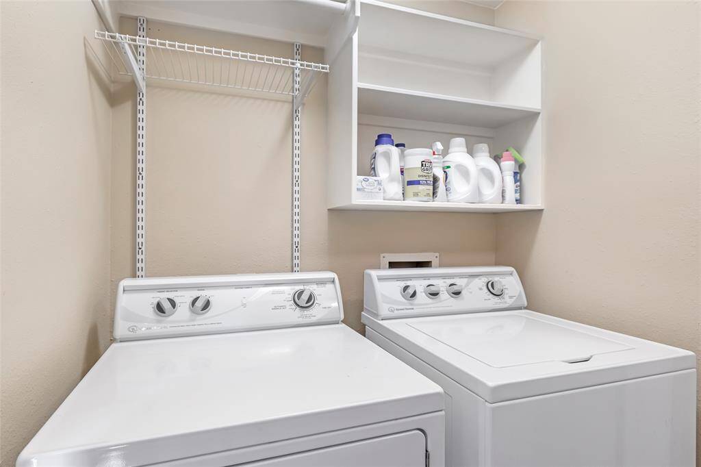 This photo shows a laundry room that fits a washer and dryer side by side. There's a shelf above for holding detergent and cleaning supplies, and an additional wire shelf for extra storage. The walls are a neutral color, providing a clean and organized space.