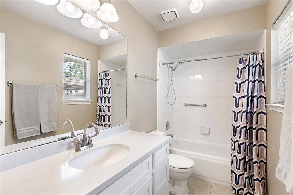 This bathroom features a clean, modern design with a white vanity and sink, a large mirror, and overhead lighting. It includes a shower-tub combo with neutral tiled flooring. Natural light enters through a window, adding brightness to the space.